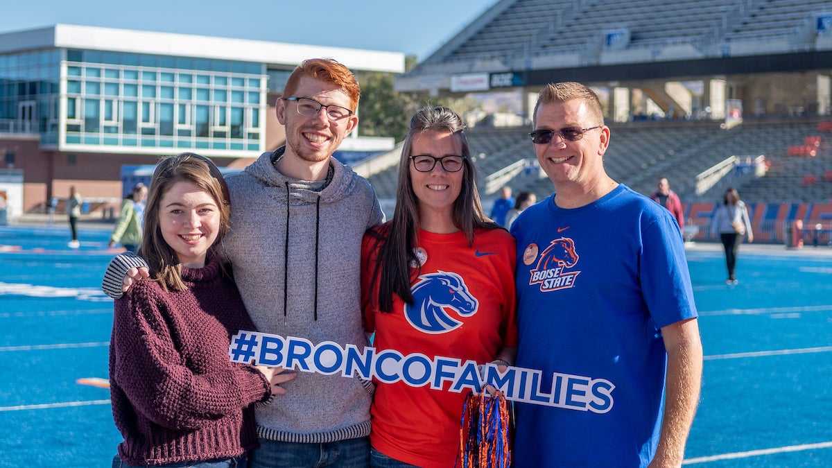 A family of four stand on the blue football turf. They are holding a Bronco Families sign while posing.