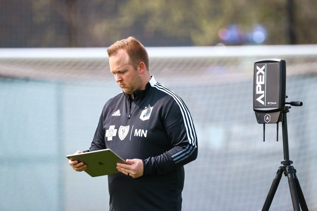 Craig Lane holding an iPad, standing on a soccer field