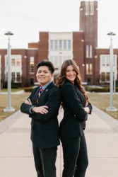 Two students standing back to back with arms folded in front of brick building