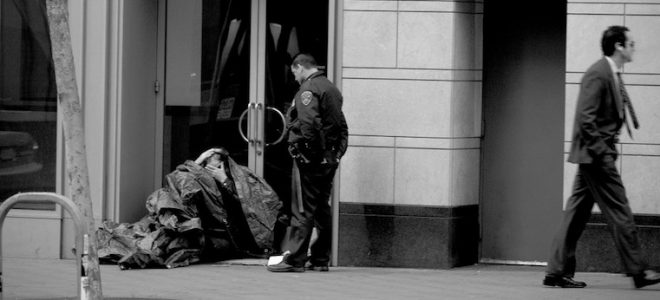 A police officer stands over a person experiencing homelessness, who is sitting in a doorway of a business covered with a tarp