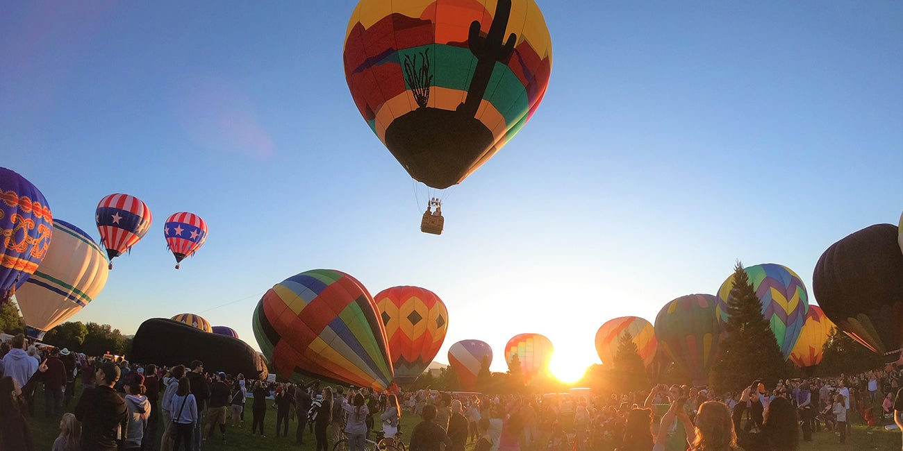 A hot air ballon hovers in the air above a field of people and hot air balloons