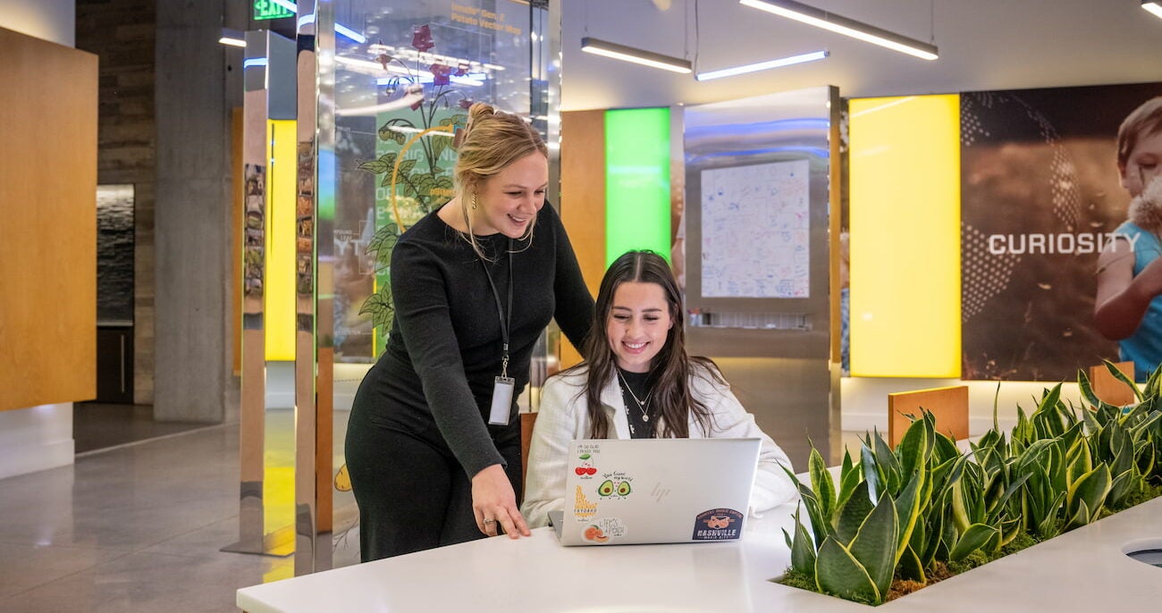 Boise State intern working at a laptop in the Simplot headquarters while her mentor looks over her shoulder and smiles