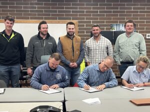 three community members sitting at a table signing paperwork while 5 other community members stand behand the table and look at the camera