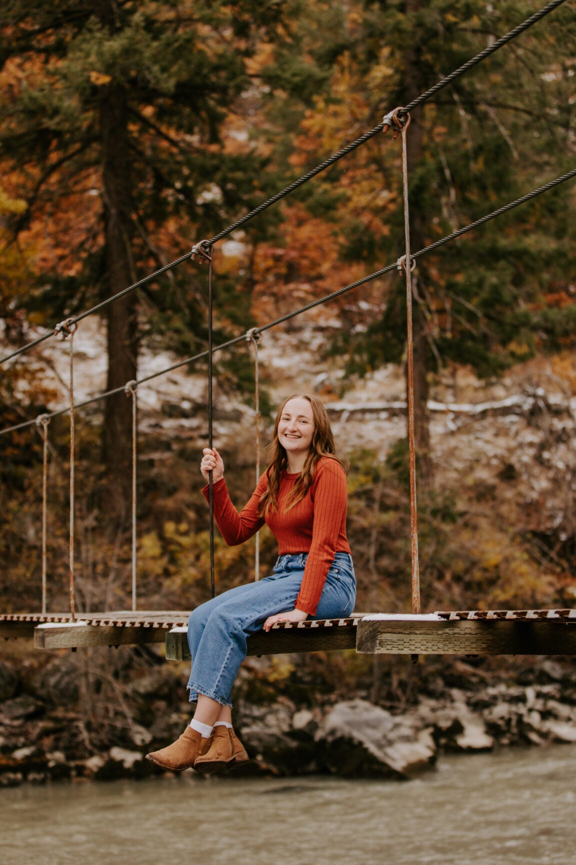 Photo of Haley sitting with her feet dangling on a rope and wood bridge over a river with trees in the background