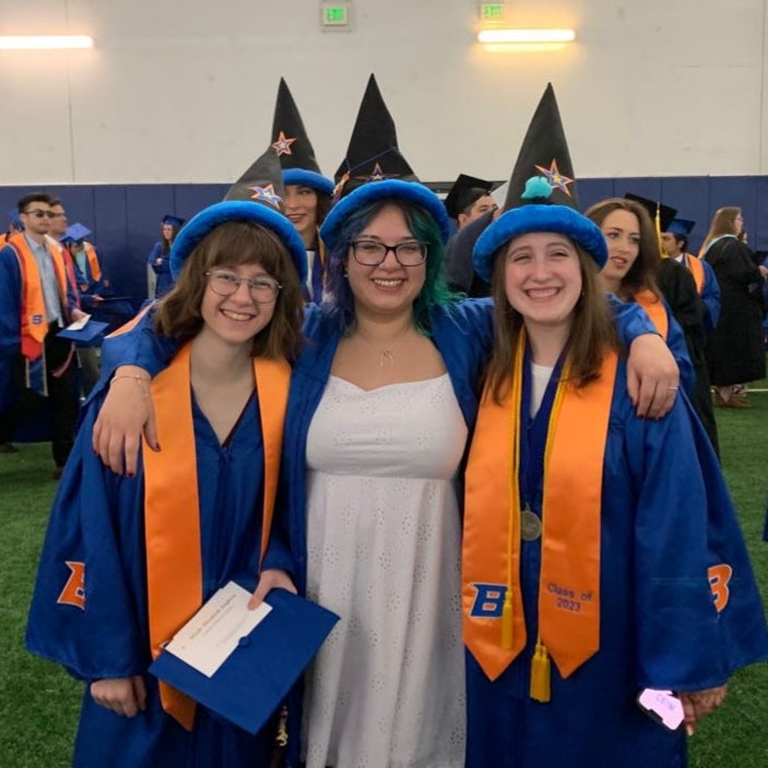 Photo of AJ Santana Cabrera standing between two female students, all wearing graduates gowns and wizard hats