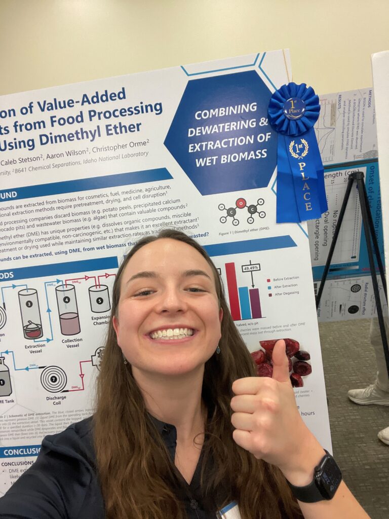 Photo of Kylie Johnson standing in front of her blue ribbon winning poster at the INL, smiling and giving a thumbs up