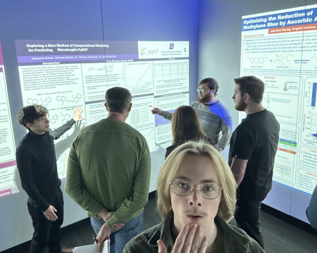 Photo of Matthew Lindley standing in front of a group of students talking to their instructor about their PChem poster