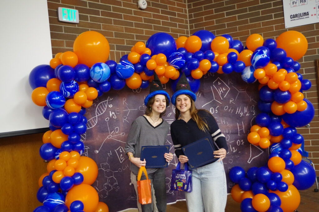 Kelso Hinrichs and Molly Gage wearing their wizard hats and holding their chemistry department graduate certificates, standing in front of a chemistry backdrop surrounded by blue and orange balloons