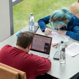 Two students are studying at a table. One uses a laptop and the other uses paper and pens.