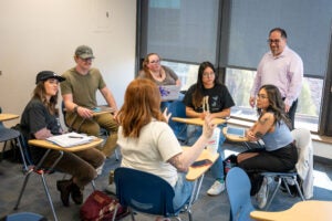 A professor stands watching as their students sit in a circle and talk.