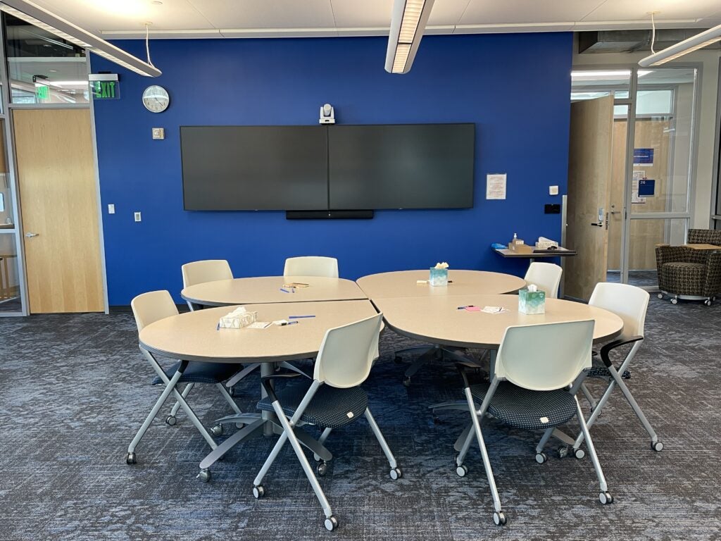 A photo of wall-mounted dual display monitors in the CTL Learning Lounge, and adjustable camera above the monitors, and the seating arrangement of four teardrop tables and eight movable chairs facing the monitors. The photo also shows extra floor space to accommodate additional seating.