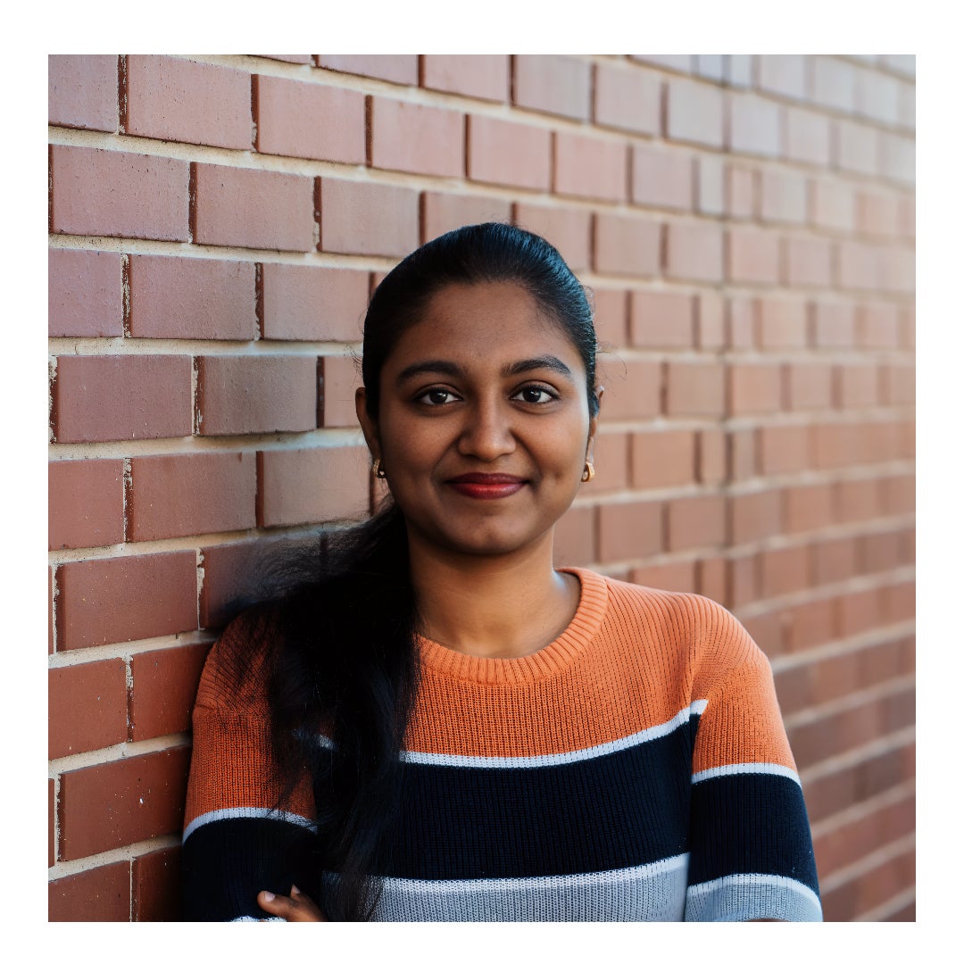 Sharadha, a woman with long brown hair, red lipstick, and gold earrings, leaning against a brick building.