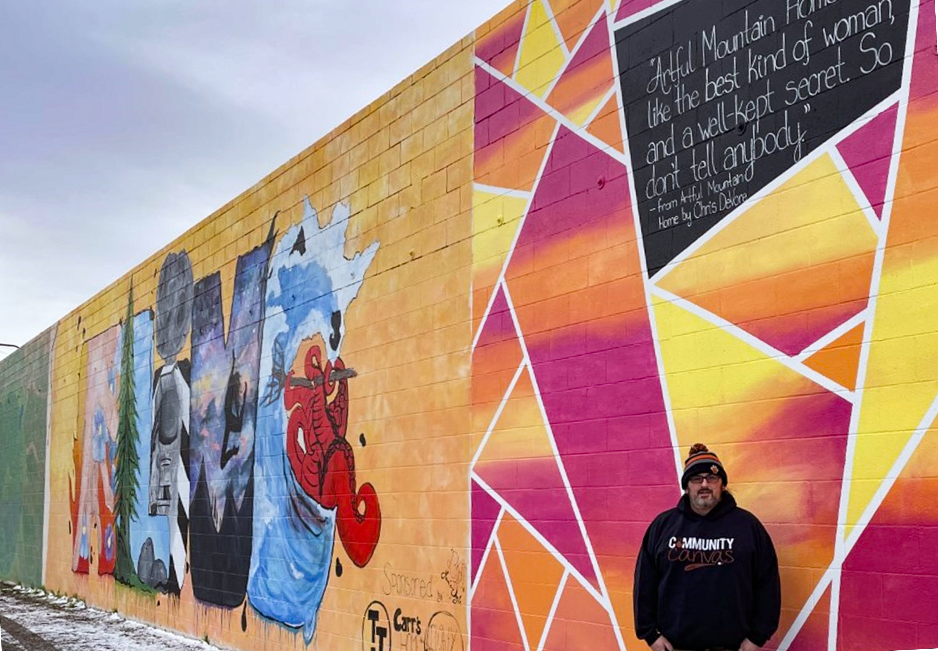 Chris DeVore stands in front of a wall of pink, orange and yellow murals.