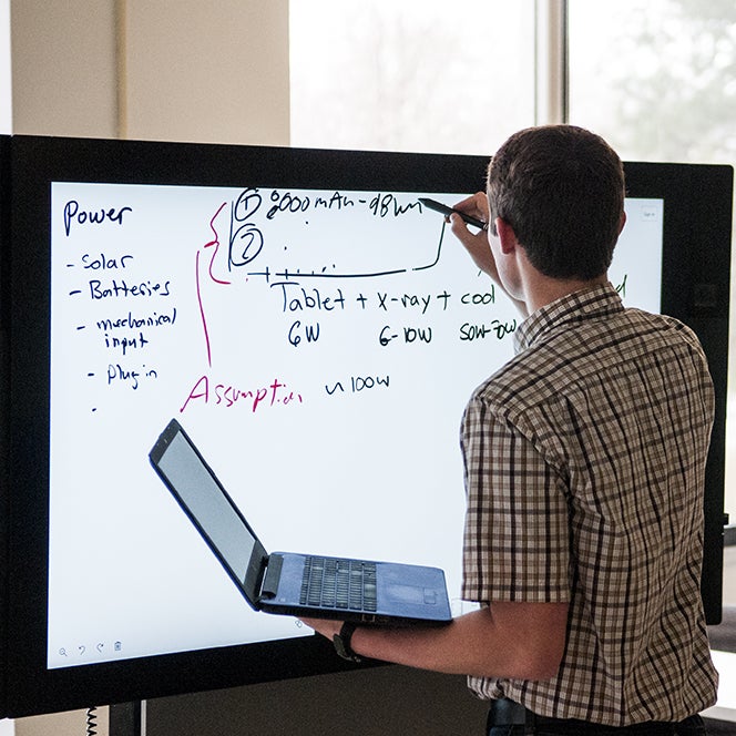 Man writing on an electronic whiteboard and holding a laptop