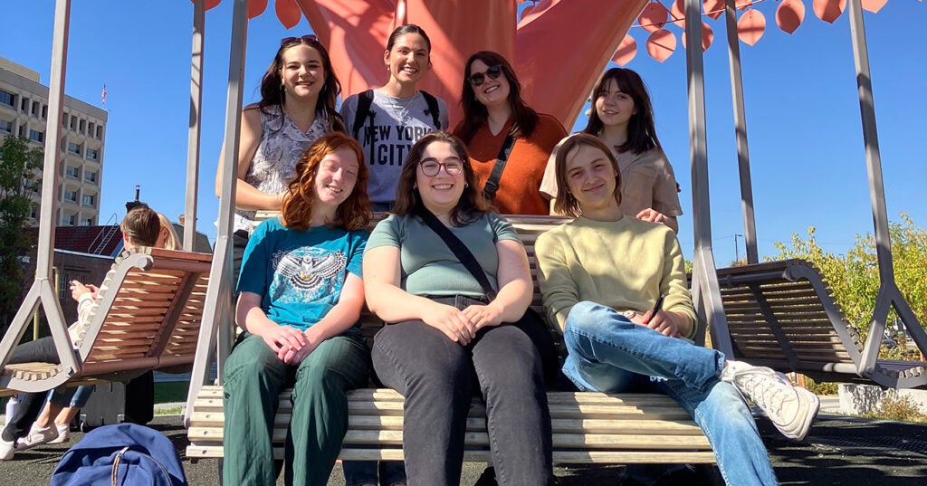  students sit in swing in front of pink metal sculpture depicting a tree.