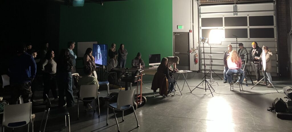 Students stand in front of a green screen while a filmed interview occurs 