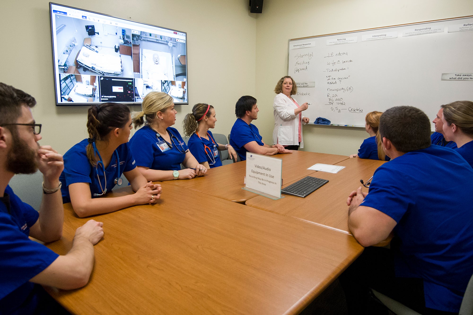 A professor wearing a white coat teaches a group of students wearing scrubs