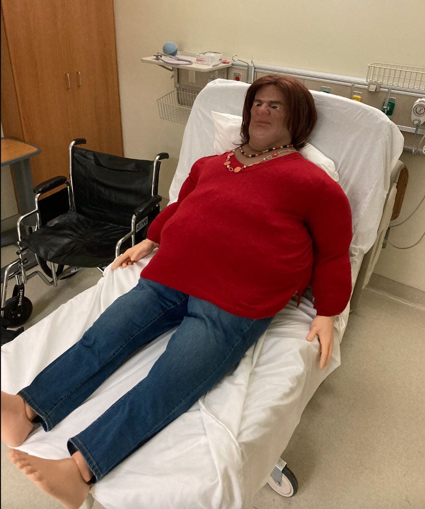 A manikin with obesity lies in a red sweater on a hospital bed.