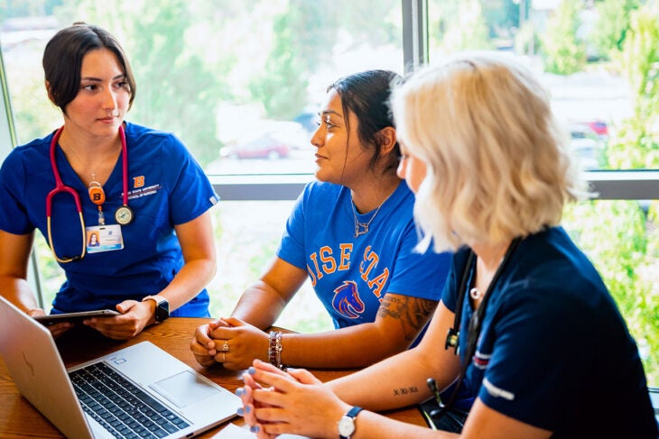 Three students sit around a table with a laptop. Two students are dressed in scrubs.