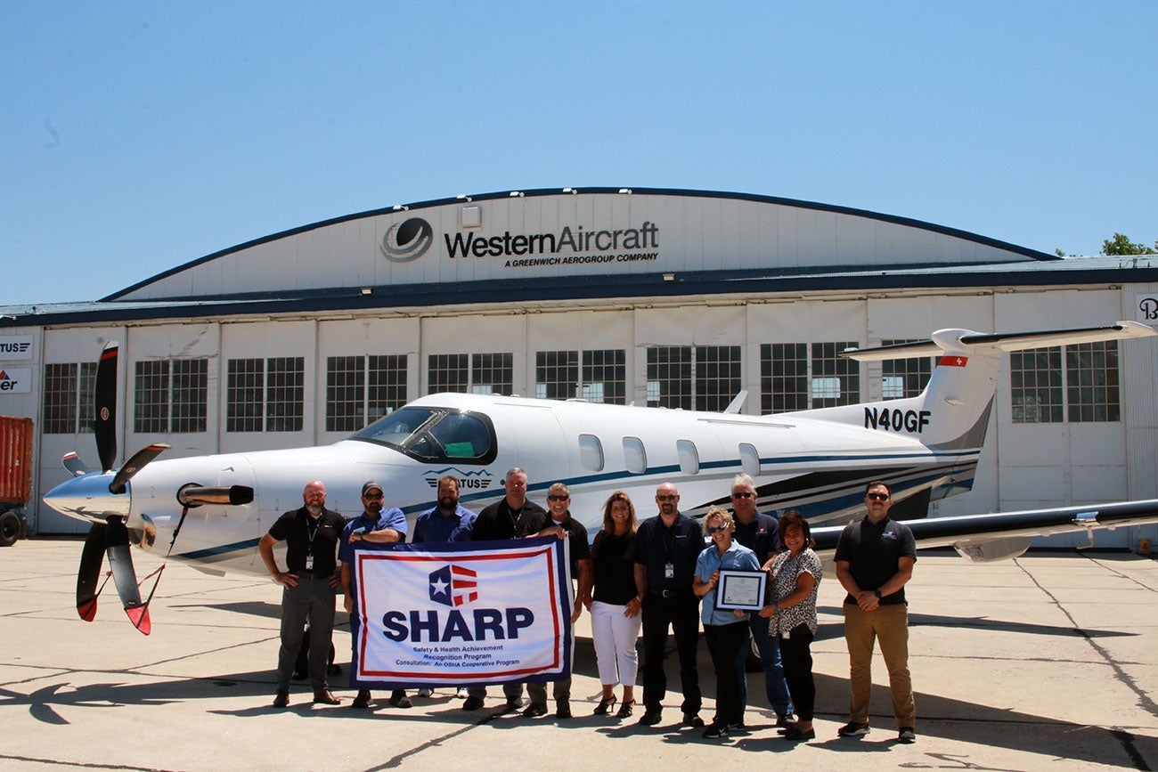 Western Aircraft employees and Idaho OSHCon team members pose with a plane in front of a Western Aircraft building while holding a SHARP flag