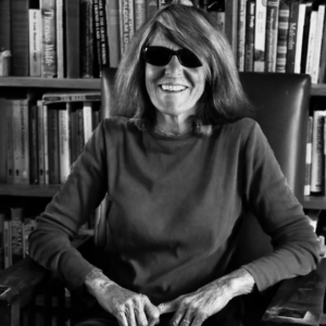 A black and white photo of writer Joy Williams wearing sunglasses, sitting in a chair, smiling at the camera. 