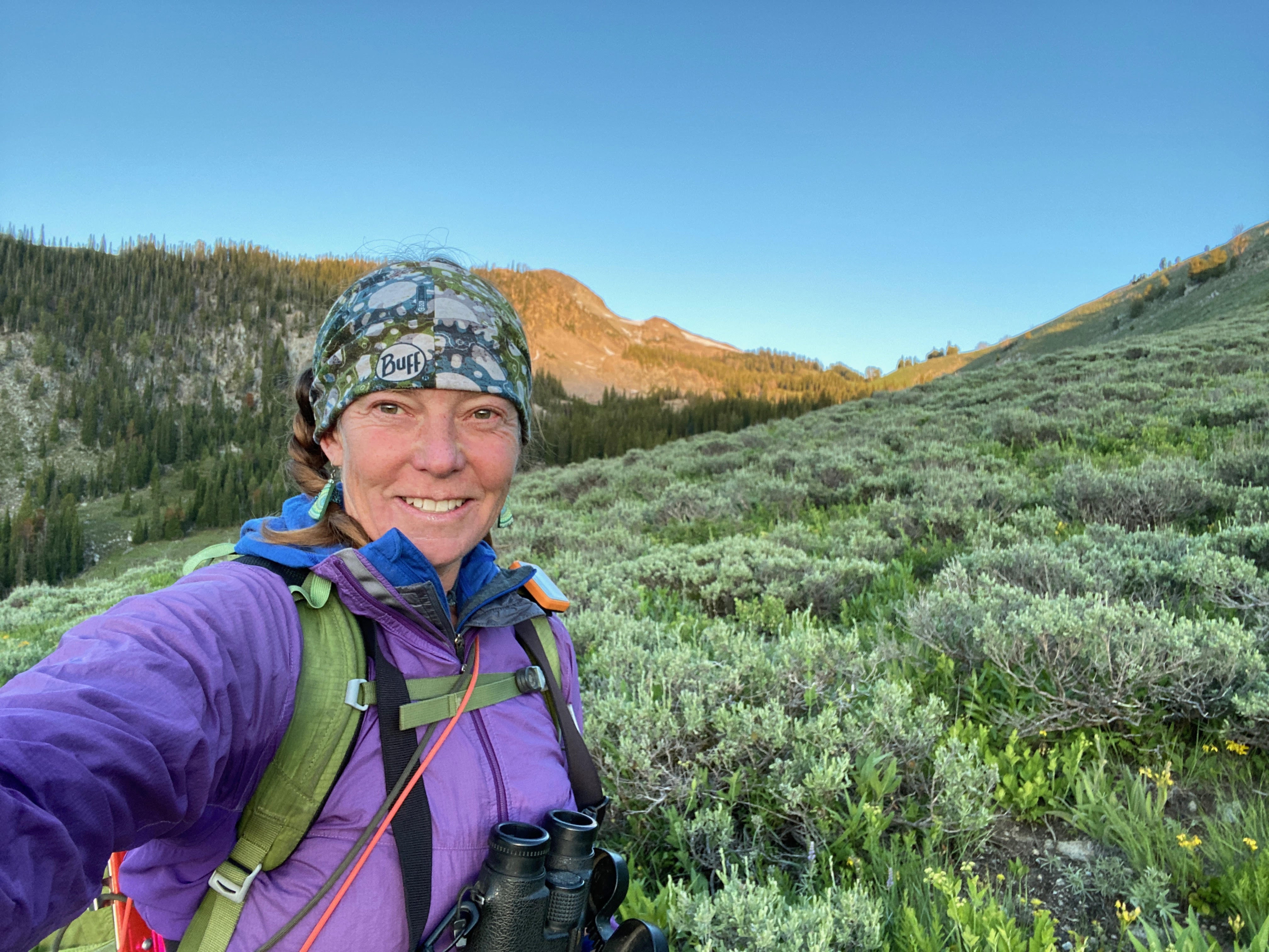 a biologist stands taking a selfie with sagebrush steppe and mountains in the background. She is smiling and wearing binoculars and a backpack