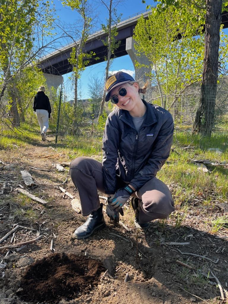 a young college student in work boots, gloves, and sunglasses, smiles at the camera while kneeling down to plant a small seedling. the highway 21 bridge and cottonwood trees of the diane moore nature center are in the background