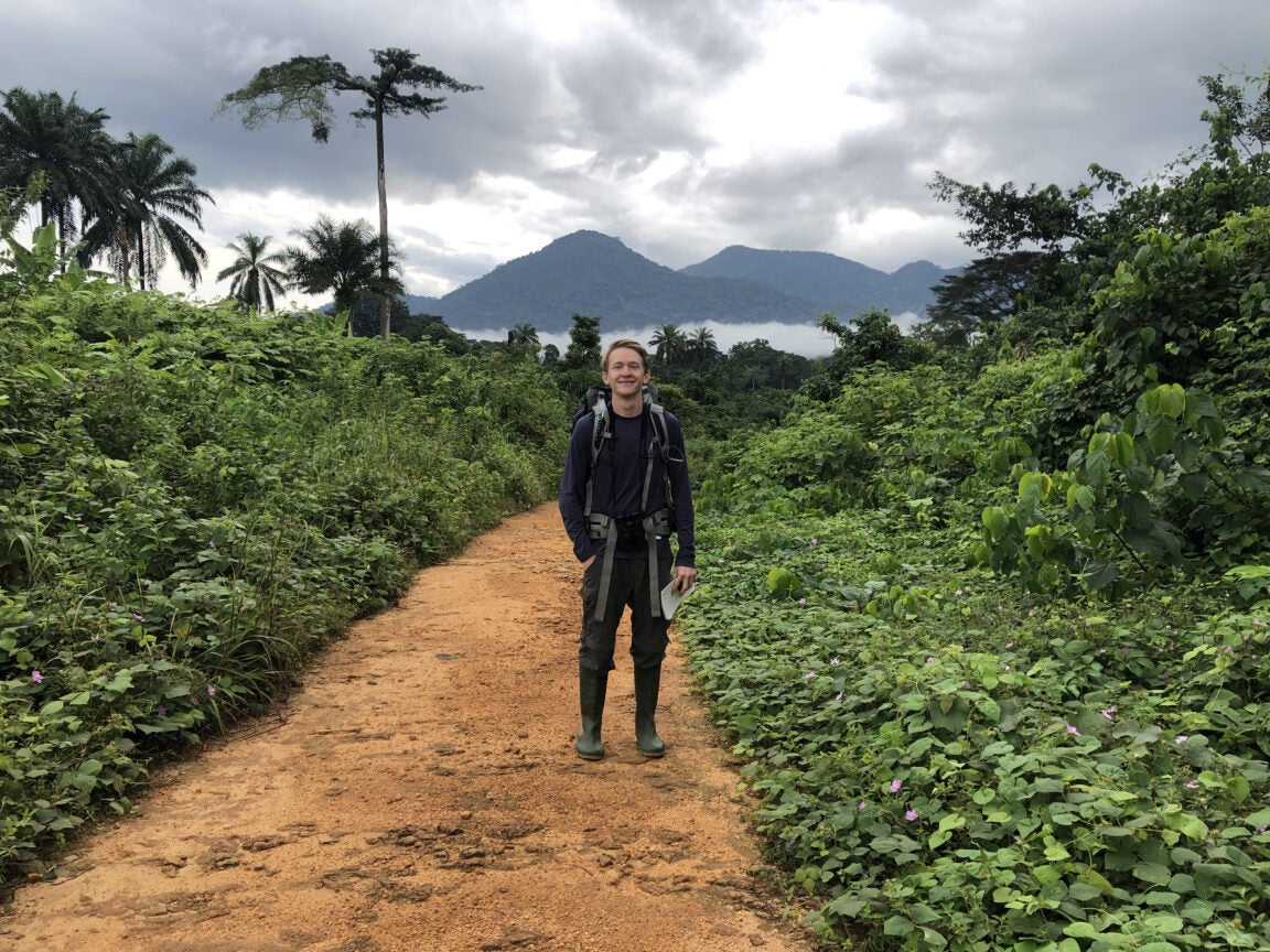 Colin stands on a reddish clay path. He's wearing mud boots and a hiking pack. Behind him is lush green vegetation and high rainforest mountains in the background