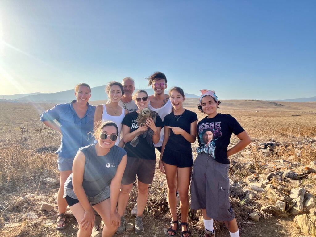 eight people stand together smiling. the person in the center is holding a black kite (a medium sized brownish hawk). It's a bright cloudless sunny day and the grasslands in the background are glowing amber brown