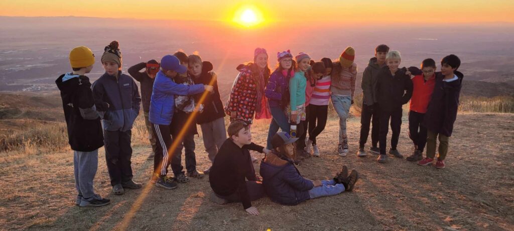 A group of 18 5th graders stands together linking arms and smiling in front of a beautiful sunset view at Lucky Peak