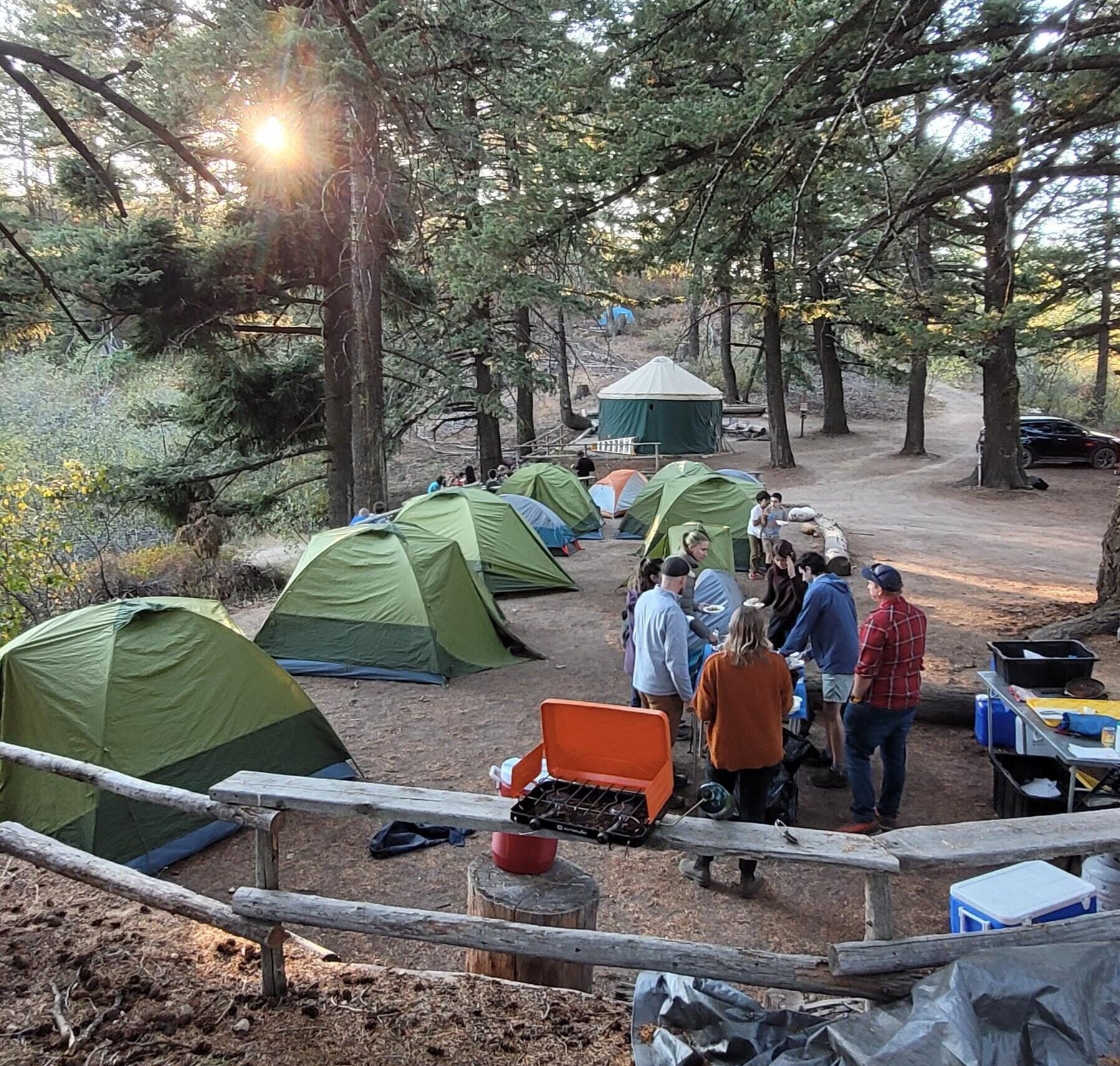 the main camping pad at Lucky Peak dotted with half a dozen green tents. A group of adults stand around tables with camp stoves and dishwashing gear. the lucky peak yurt and douglas fir trees are in the background