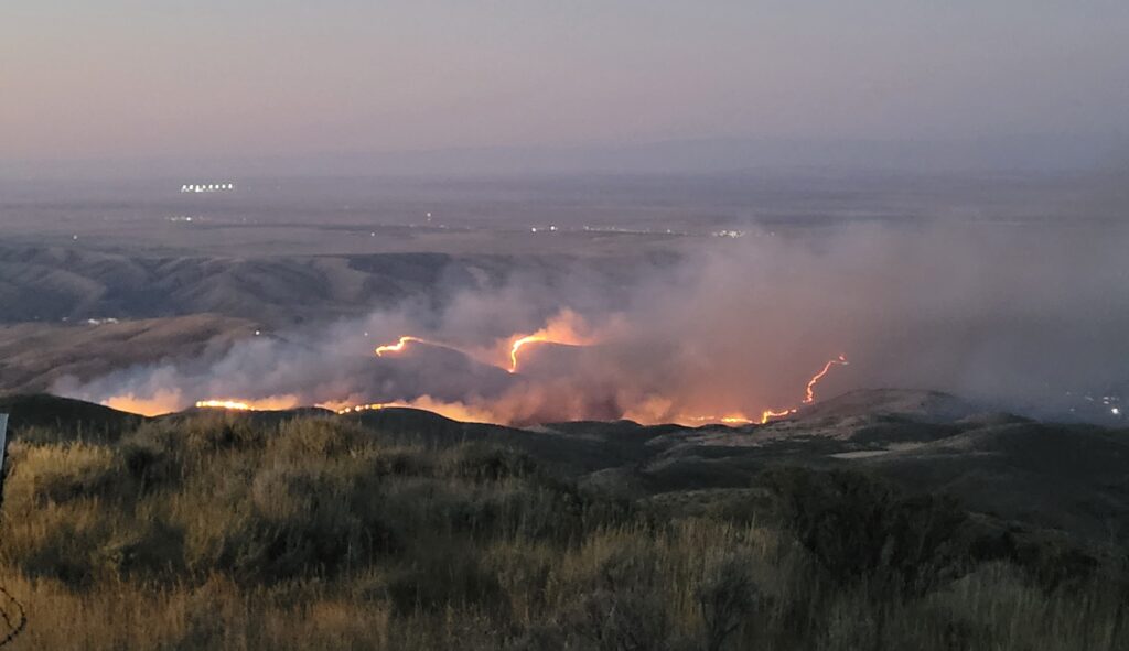 two glowing orange lines of fire border a charred black landscape. the early morning light is purplish. billows of smoke are rising from the fire area. the fire looks quite close