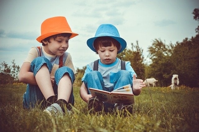 Two kids reading in a meadow and wearing colorful hats.