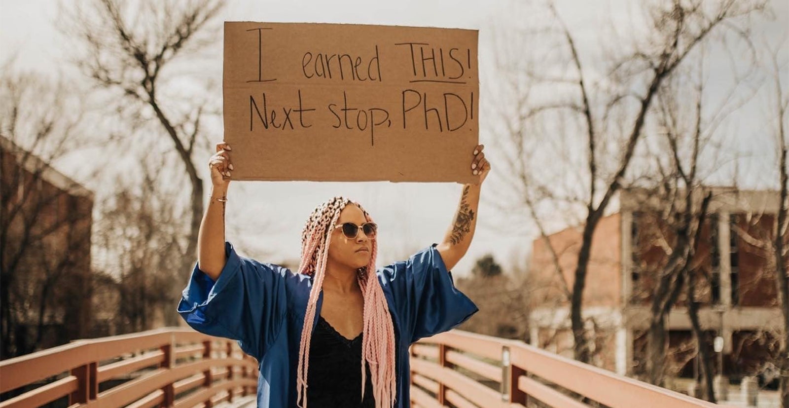 Student wearing graduation regalia stands on bridge holding sign 