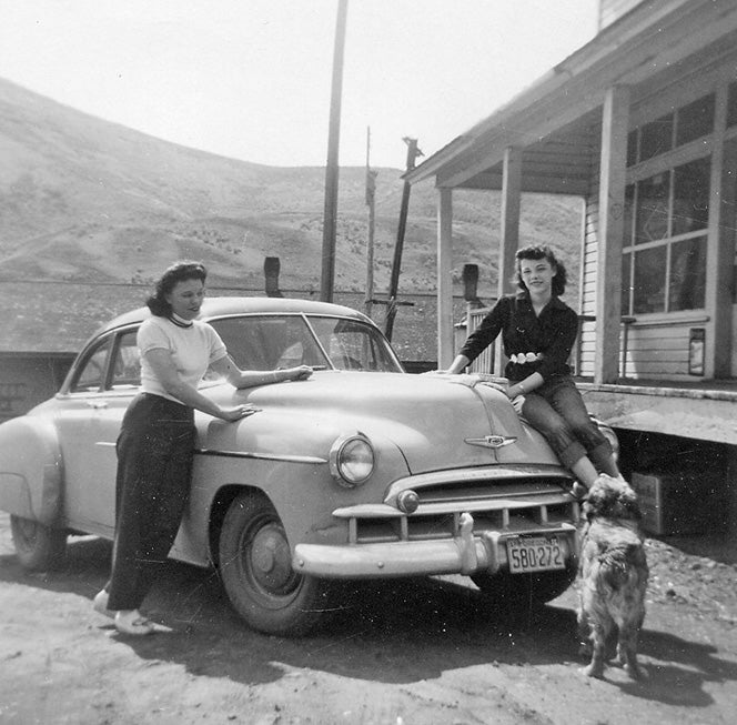 Two women lean on a car, a dog looks up at them