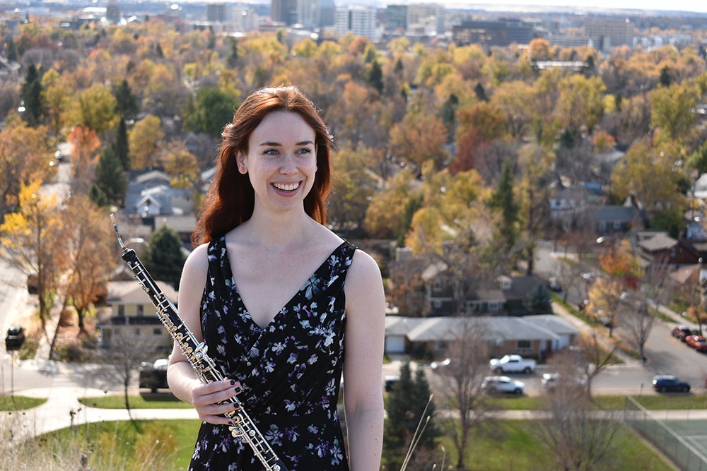 Photo of Rachel standing outside holding an oboe. Behind her are residential home and autumn colored trees