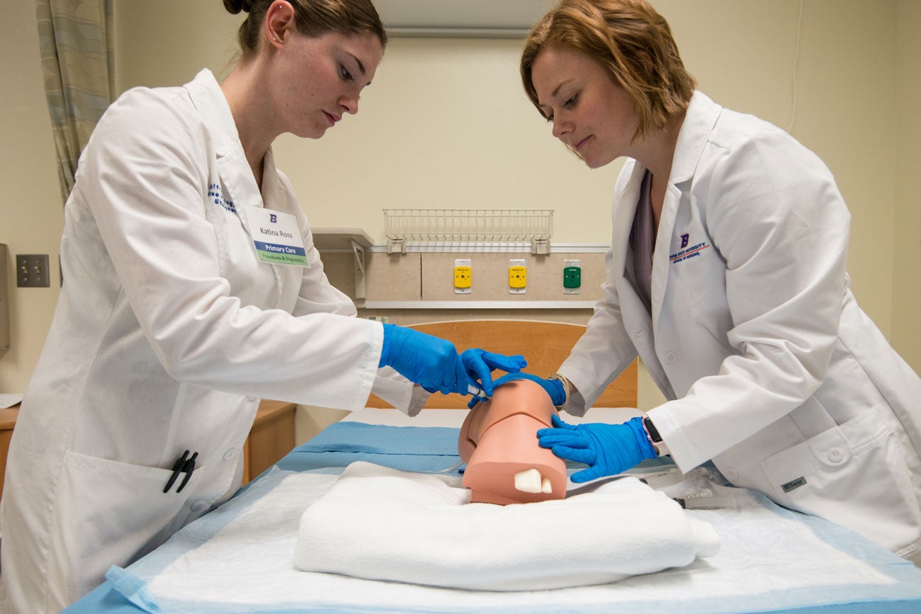 Two nurse practitioner students practice injections on a manikin leg joint.