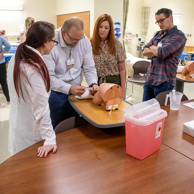 Three nurse practitioner students gather around a table as a faculty supervises their joint injection practice on a manikin.