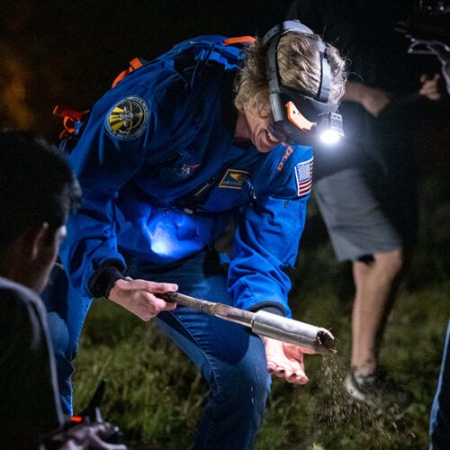 Woman in blue Nasa jacket wearing a headset inspecting a soil sample at the end of a collecting rod.