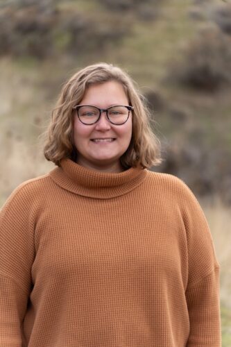 Young woman with blonde hair and glasses in brown turtleneck stands outside