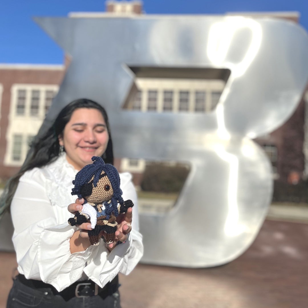 Student hold crocheted doll in front of B statue