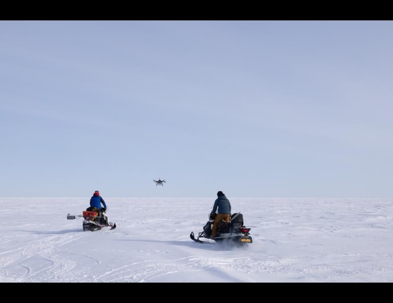 two men on snowmobiles ride across frozen sea ice towards a distant flying drone