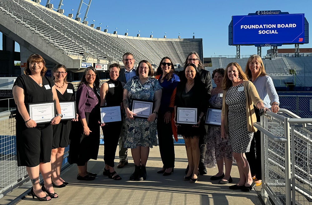 A group of 12 faculty and staff pose outside in Albertsons Stadium holding framed awards from the University Foundation.