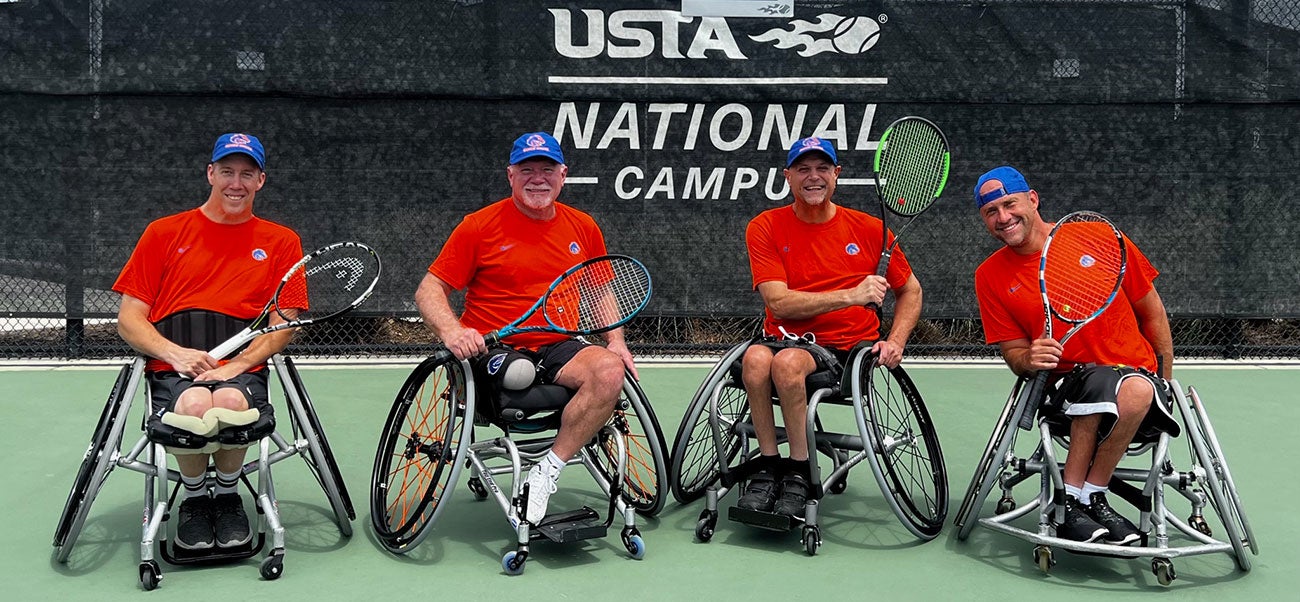 Four men in Bronco gear in wheelchairs on tennis court holding tennis rackets