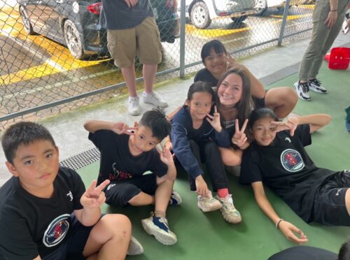 Elle, a US college student sits on a sports court surrounded by Taiwanese elementary schoolers who are smiling and giving the peace sign.