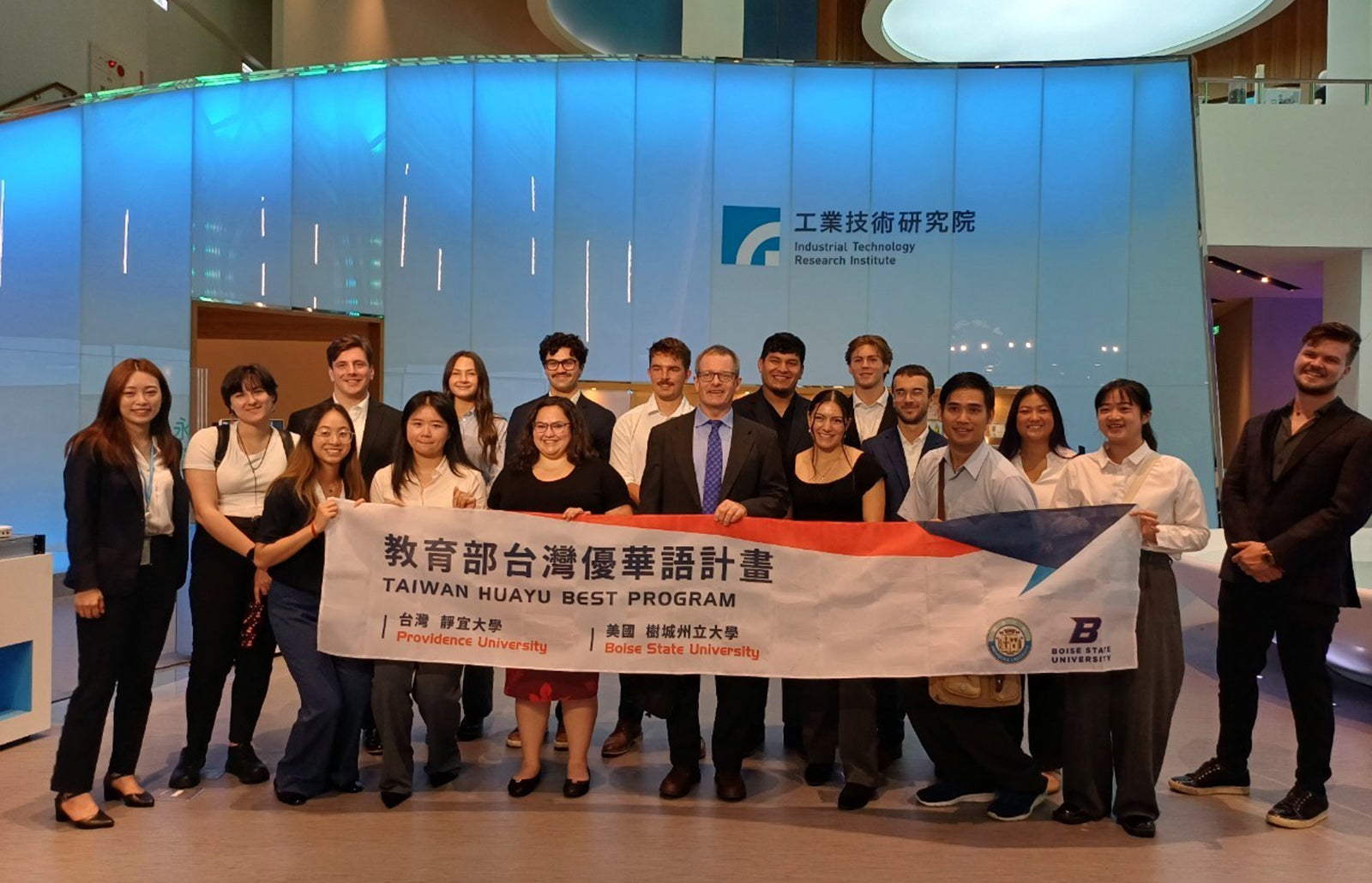 A group of students stands in in front of a lit wall with the Industrial Technology Research Institute logo on it. The group is holding a banner that says "Taiwan Huayu Best Program" with Boise State and Providence University logos on it.