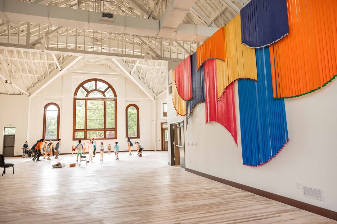 In a large bright room with colorful wall hangings, a group of children and instructors engage in a group activity.