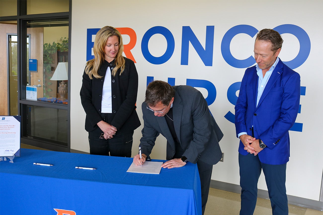 Two people look on as the dean of the Episcopal Cathedral signs a document on a table with a blue tablecloth.