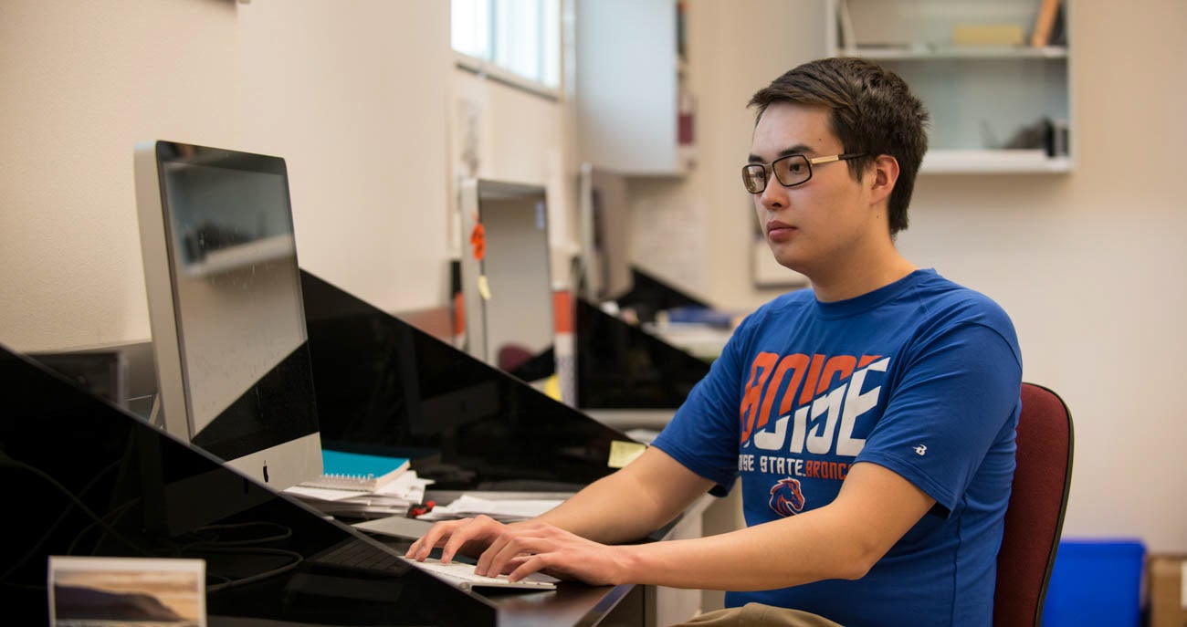 A man sits at a desk while working on a computer wearing a Boise State Broncos shirt.