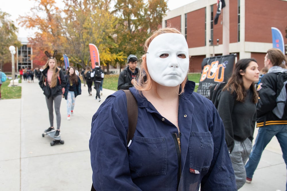 a student in a full blue jumpsuit wears a white full face mask. She is surrounded by dozens of other students who are walking on campus, some also wearing Halloween costumes.